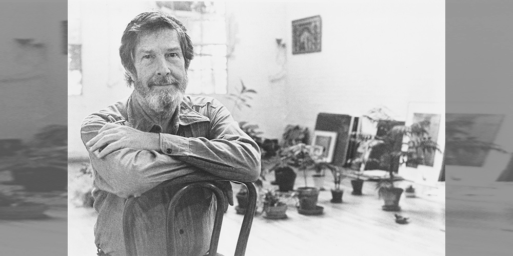 John Cage sitting in a chair in a large room with many plants